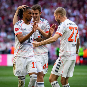 Three soccer players in Bayern Munich jerseys celebrate a goal on the field. They express excitement, with one player raising his arms and another smiling broadly. The background features a cheering crowd, enhancing the moment of victory and team camaraderie.