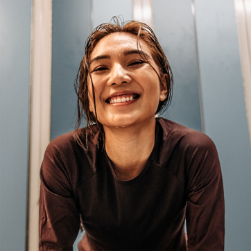 A smiling young woman with wet hair leans forward, showcasing her joyful expression. She wears a dark athletic shirt, and the background features light blue walls, creating a vibrant, energetic atmosphere. The image conveys a sense of happiness and vitality, likely after physical activity or exercise.