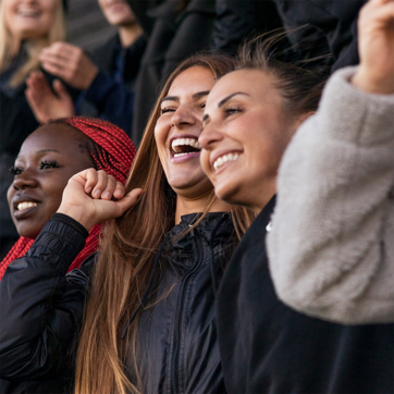 Three young women are smiling and cheering enthusiastically. They appear to be celebrating or enjoying a moment together, dressed in casual jackets. The atmosphere is lively, with a joyful expression shared among them.
